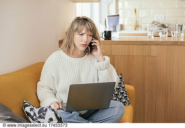 Dissatisfied woman talking on smartphone  sitting on couch with laptop