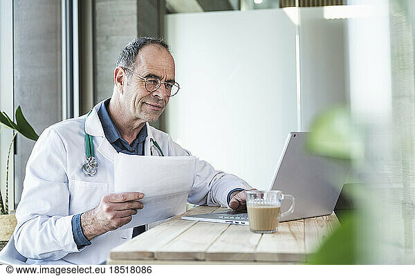 Doctor holding medical report and using laptop at desk
