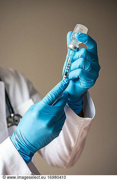 Doctor in white coat and gloves drawing vaccine into a syringe.