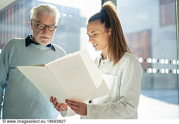 Doctor reading file by senior patient at sunny day in hospital