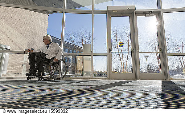 Doctor with muscular dystrophy in wheelchair at hospital entrance pressing accessible door plate