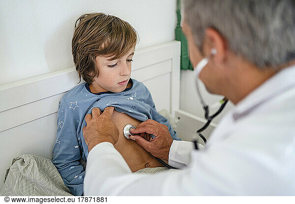 Doctor with stethoscope examining boy in bed at home