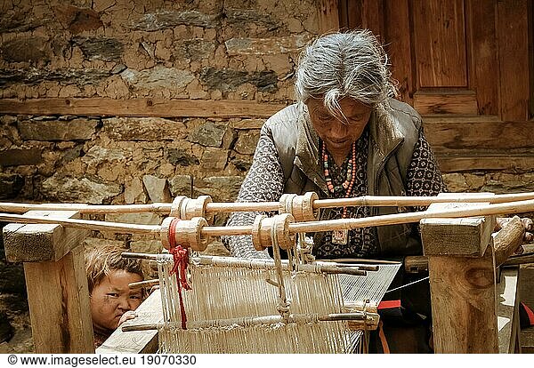 Dolpo  Nepal  circa June 2012: Old grey-haired woman dressed in grey vest with necklaces made of beads looks down and weaves on simple wooden loom in Dolpo  Nepal. Documentary editorial  Asia