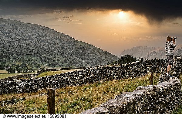 Dramatischer Sonnenuntergang mit einem Reisefotografen  der auf einer Trockenmauer steht und den Kirkstone Pass  Lake District National Park  Cumbria  England  fotografiert. Der Nationalpark ist ein UNESCO-Weltkulturerbe