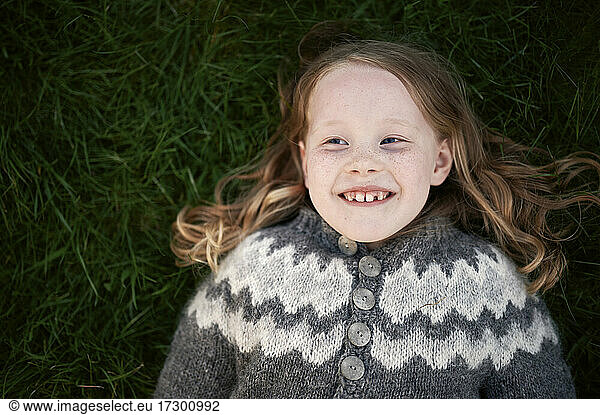 Draufsicht auf ein entzücktes Mädchen in Strickjacke  das lächelnd auf einer grünen Wiese liegt und wegschaut