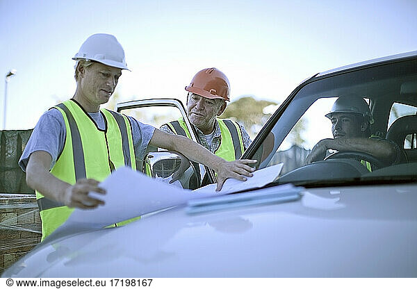 Drei Bauarbeiter unterhalten sich über Pläne  die auf der Motorhaube eines Autos ausgelegt sind