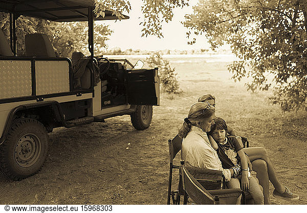 Drei Generationen einer Familie sitzen in Klappstühlen in einem Safarijeep  unter Bäumen  einfarbig.