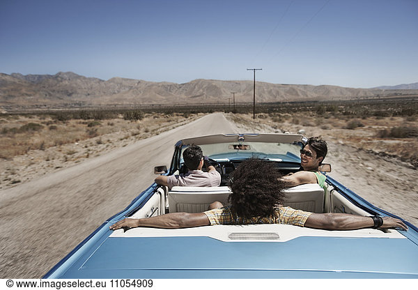Drei junge Leute in einem hellblauen Cabriolet,  die auf offener Straße über eine flache,  trockene Ebene fahren