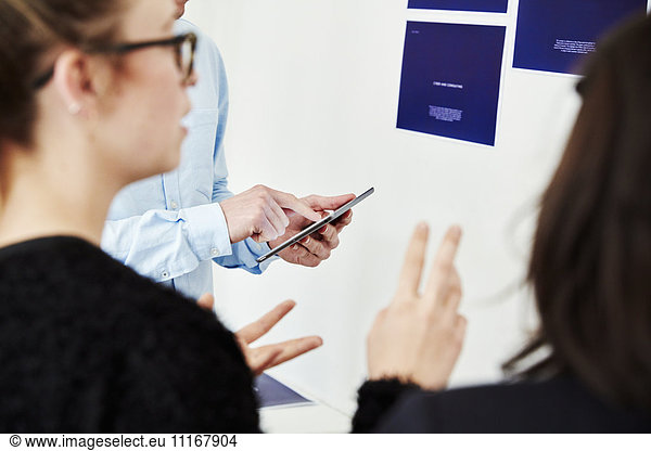 Drei Personen benutzen ein Smartphone neben einer mit Plakaten bedeckten Wand.