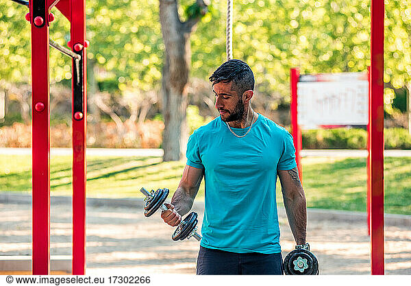 Dunkelhaariger Athlet mit Bart trainiert mit Hanteln in einem Park. Outdoor-Fitness-Konzept.