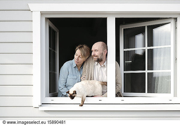 Ehepaar mit Katze  die sich aus dem Fenster ihres Hauses lehnt