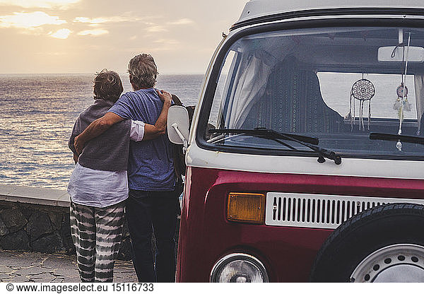 Ein älteres Ehepaar reist in einem Oldtimer-Van und schaut mit den Armen auf das Meer