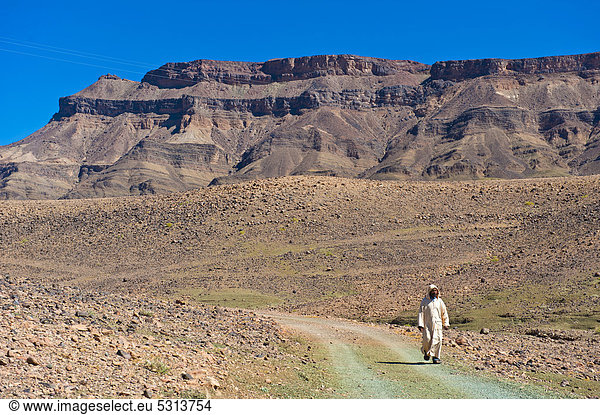 Ein Berber in traditioneller Djellabah geht auf einem Fahrweg  Djebel Kissane dahinter  Draa-Tal  Südmarokko  Marokko  Afrika