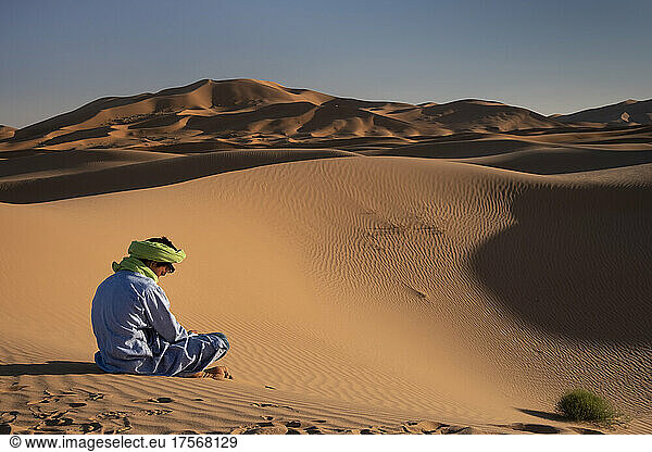 Ein Berber in traditioneller Kleidung sitzt inmitten der Sanddünen von Erg Chebbi  Sahara-Wüste  Marokko  Nordafrika  Afrika