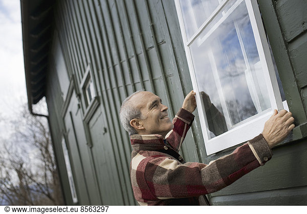 Ein Bio-Bauernhof im Winter. Instandhaltung. Ein Mann hält ein Sturmfensterpaneel gegen den Fensterrahmen.