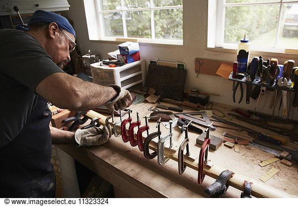 Ein Bogenbauer  der in seiner Werkstatt an einem Holzbogen arbeitet und das Holz durch Festhalten in Klammern formt.