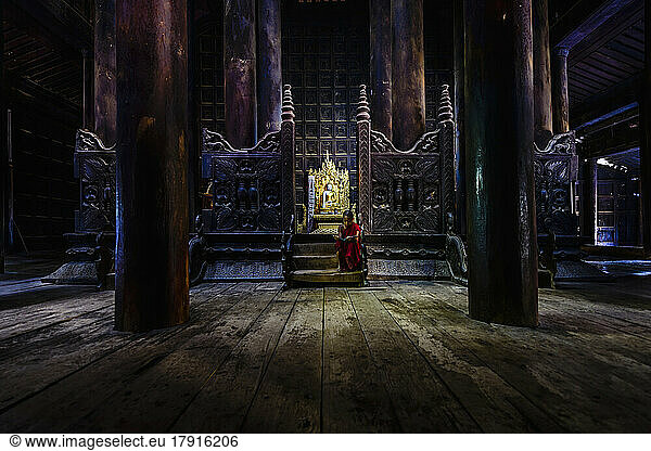Ein buddhistischer Tempel mit hölzernen Säulen und geschnitzten Schirmen  einem goldenen Thron und einem jungen Mönch  der auf den Stufen sitzt.