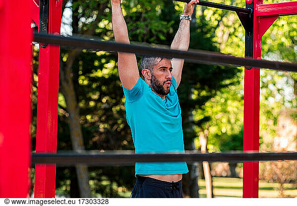 Ein dunkelhaariger Athlet mit Bart hängt an einer Calisthenics-Stange. Blick zwischen die Stangen. Outdoor-Crossfit-Konzept.