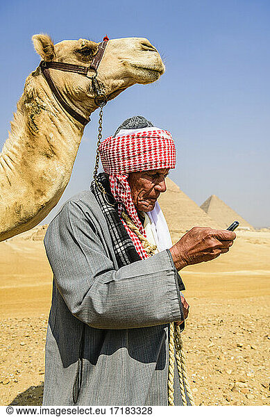 Ein Führer in traditioneller arabischer Kleidung  Gewand und Turban  der ein Mobiltelefon benutzt  und sein Kamel.
