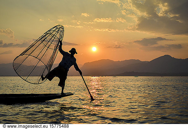 Ein Fischer  der auf einem Bein auf einem Boot balanciert  mit einem großen Fischerkorb in der Hand  fischt auf traditionelle Weise auf dem Inle-See bei Sonnenuntergang in Myanmar.