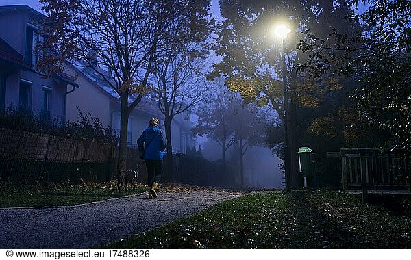 Ein Frau geht an einem dunklen und kalten Herbstmorgen mit ihrem Hund durch eine einsame Parkanlage in Markt Schwaben  Bayern  Deutschland  Europa