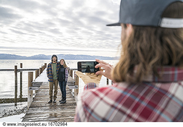 Ein Freund macht ein Handyfoto für ein Paar  das am Ende eines Piers steht