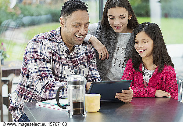 Ein glücklicher Vater und seine Töchter benutzen ein digitales Tablet in der morgendlichen Küche