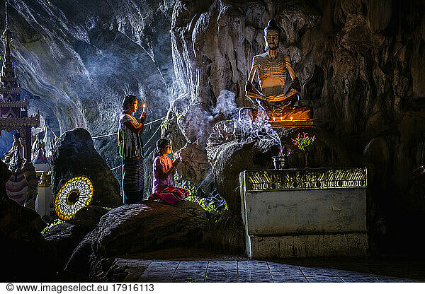 Ein Höhlentempel mit Buddha-Statuen  brennenden Kerzen und zwei knienden  betenden Mädchen.