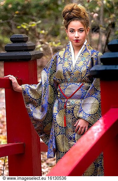 Ein 14-jähriges brünettes Mädchen in einem Kleid im asiatischen Stil schaut in die Kamera und steht auf einer roten japanischen Fußgängerbrücke.