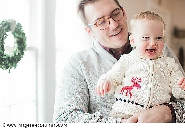 Ein 30-35 Jahre alter Vater lächelt  während er seinen kleinen Sohn zu Weihnachten im Arm hält.