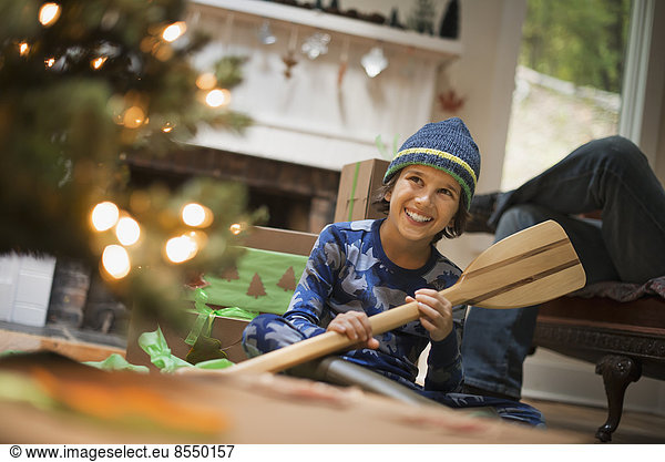 Ein Junge an einem Weihnachtsbaum  der ein Geschenk auspackt  ein hölzernes Ruder.
