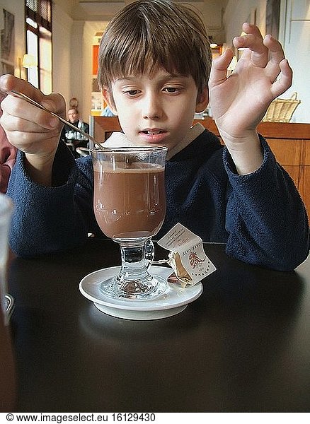 Ein Junge probiert ein schaumiges heißes Schokoladengetränk in einem Glas mit Schokoladensüßigkeiten und signalisiert mit seinen Händen Vollkommenheit im sonnenbeschienenen Café des Cinquantenaire-Museums  Königliche Museen für Kunst und Geschichte  Brüssel  Belgien.