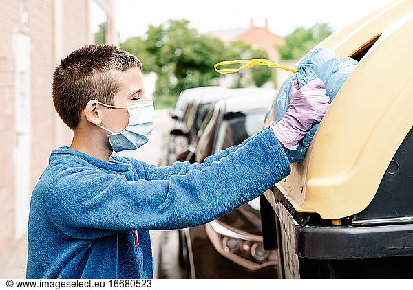 Ein Junge wirft einen blauen Müllsack in einen gelben Recycling-Container auf der Straße. Das Kind mit Gesichtsmaske und Handschuhen schiebt den Müllsack  weil er zu groß ist  um in die Tonne zu passen. horizontal