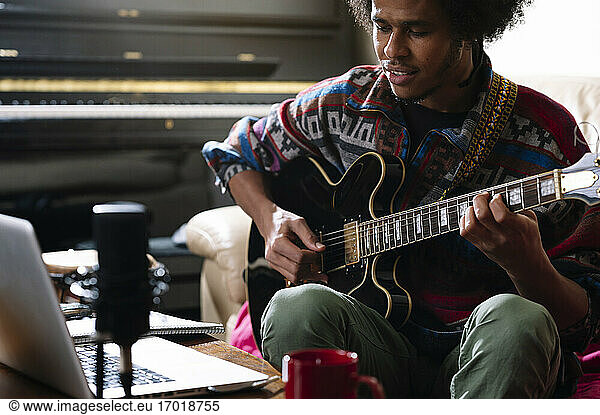 Ein junger Afroamerikaner lernt im Wohnzimmer über einen Laptop Gitarre spielen