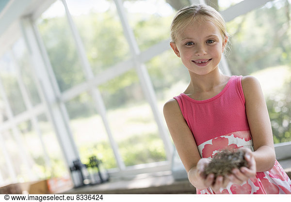 Ein junges Mädchen in einer Küche  das ein rosa Kleid trägt. Sie hält ein Vogelnest in der Hand.