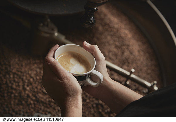 Ein Kaffeehaus. Eine Person  die eine frisch gebrühte Tasse über eine Metalltrommel mit gerösteten Kaffeebohnen hält.