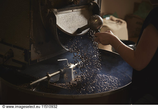 Ein Kaffeehaus. Eine Person öffnet eine Metallrinne und Kaffeebohnen fallen in eine Rösttrommel.