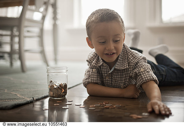 Ein Kind  das auf dem Bauch auf dem Boden liegt  mit Münzen spielt und sie in ein Glasgefäß legt.
