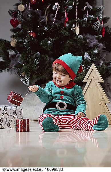 Ein kleiner 12 Monate alter Weihnachtself mit einem Weihnachtsbaum und Geschenken