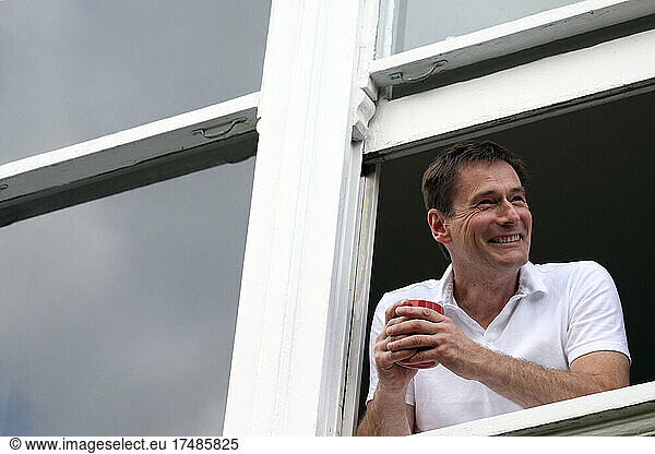Ein lächelnder Mann lehnt sich mit einem heißen Getränk aus dem Fenster.