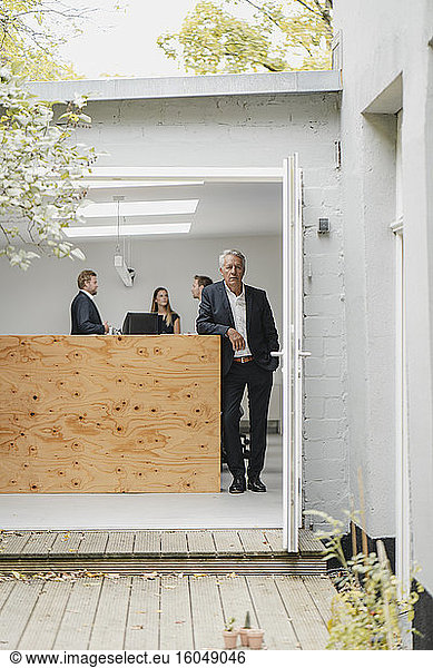 Ein älterer Geschäftsmann steht in einer offenen Bürotür  im Hintergrund arbeiten Menschen