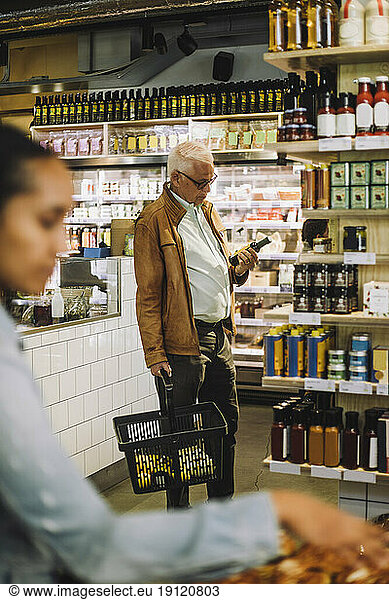 Ein älterer Mann steht mit einem Einkaufskorb und einer Flasche im Supermarkt