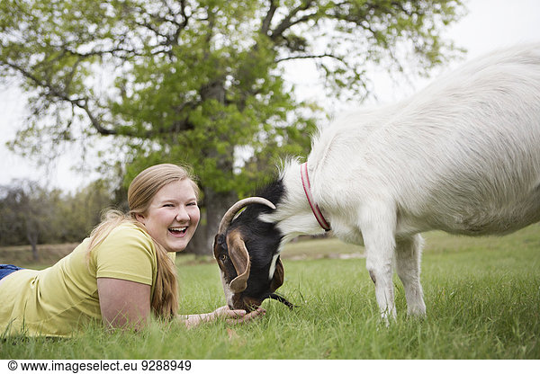 Ein Mädchen  das Kopf an Kopf mit einer Ziege auf Gras liegt.