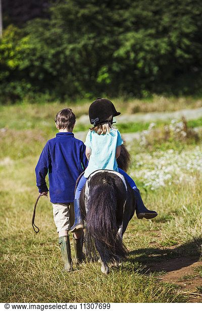 Ein Mädchen reitet auf einem Pony und ein Junge geht durch ein Feld.