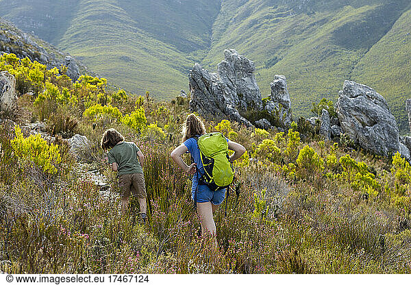 Ein Mädchen und ein Junge wandern auf einem Pfad durch Vegetation und Felsen im Fynbos