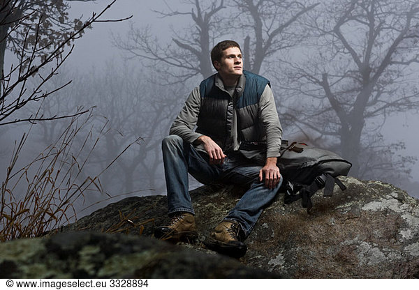 Ein männlicher Wanderer sitzt auf einem Felsen in einem nebligen Wald.
