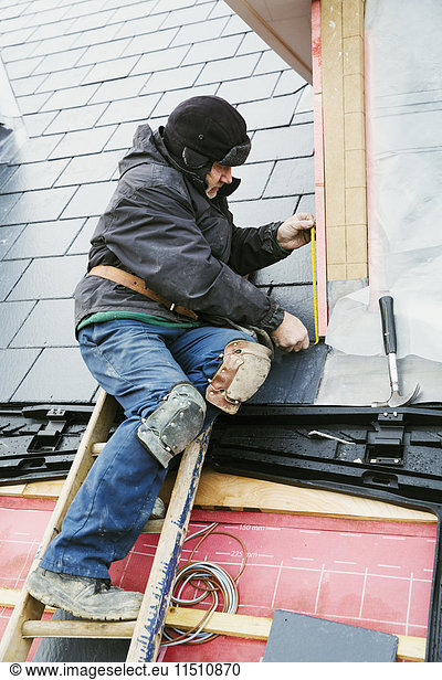 Ein Mann an der Spitze einer Leiter auf einem Hausdach  der Ziegel auf einem Gaubendach befestigt.