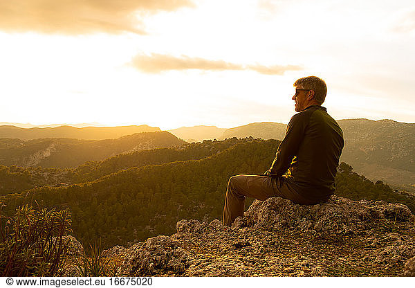 Ein Mann bei Sonnenuntergang auf einem Felsen sitzend mit Blick auf die Berge bei Sonnenuntergang