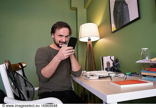 Ein Mann überprüft sein Smartphone und lächelt  als er in die Kamera in seinem Zimmer mit Büchern schaut.