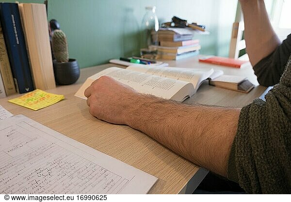 Ein Mann  der in seinem Arbeitszimmer ein Buch liest und seinen Arm in der Quarantänezeit auf den Schreibtisch legt.